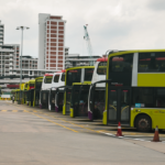 Line of double-decker buses parked at an urban terminal with city buildings in the background.