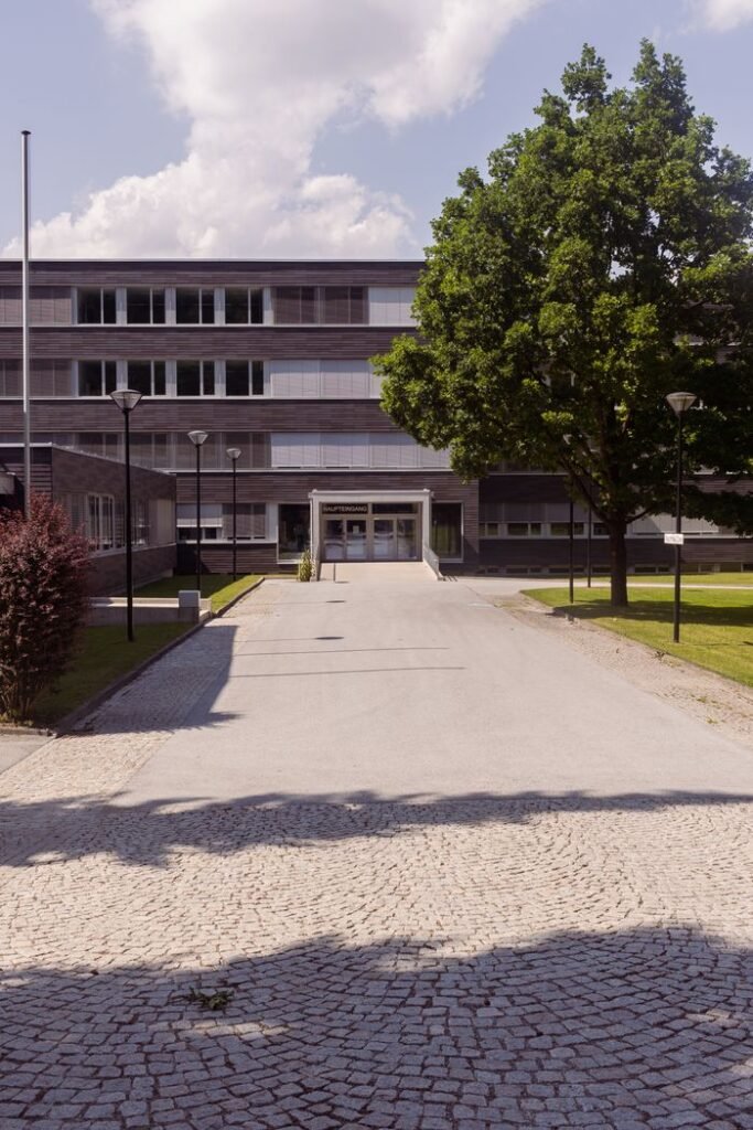 pexels-photo-18535001-18535001 A modern building with a cobblestone path, surrounded by trees on a sunny day.