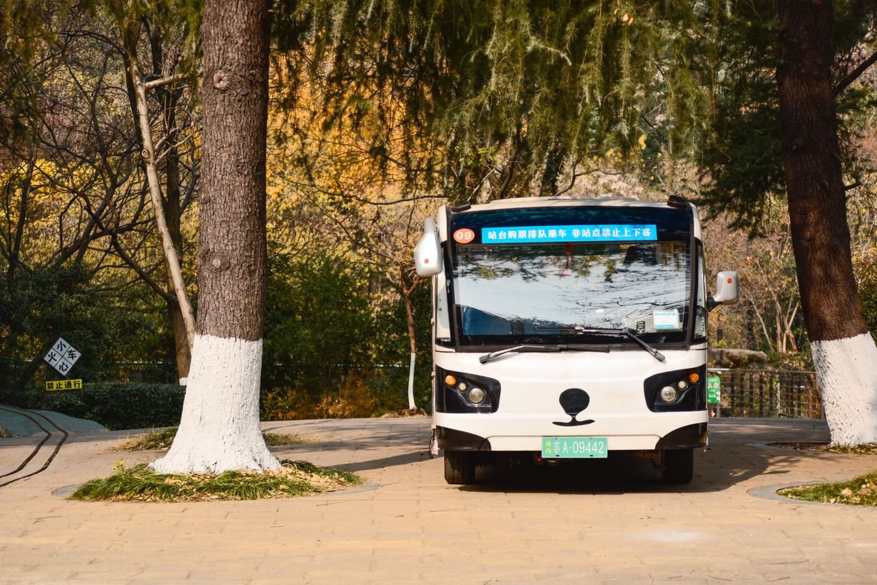 Electric shuttle bus driving on a tree-lined path in a park with autumn foliage.