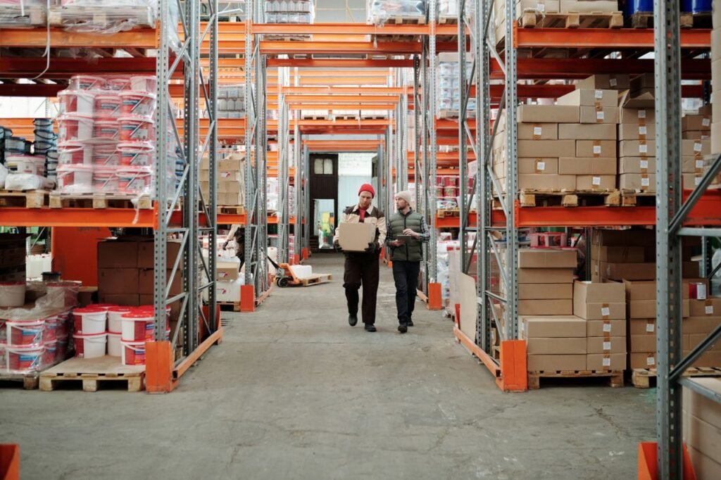 pexels-photo-4487383-4487383 Two workers handle a package in a spacious warehouse surrounded by shelves stocked with boxes and products.