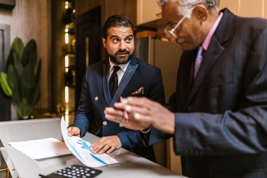 Planned Implementation Two men in suits discuss financial documents with graphs indoors.